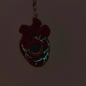 May include: A red plush heart-shaped keychain with a glowing green eye and veins. The keychain is hanging from a black string.
