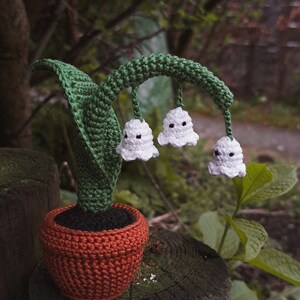 May include: A crocheted lily of the valley plant in a terracotta pot. The plant has white flowers with black dots and green leaves.