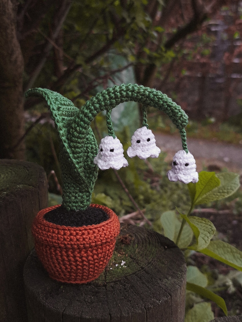 May include: A crocheted lily of the valley plant in a terracotta pot. The plant has white flowers with black dots and green leaves.