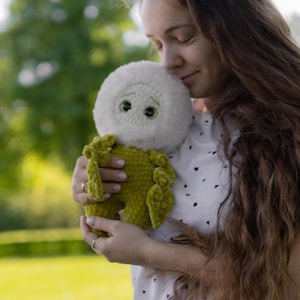 Pode incluir: Uma boneca de crochê feita à mão com cabeça branca e corpo verde-oliva. A boneca tem dois olhos pretos e está sendo segurada. A cabeça da boneca é fofa e o corpo tem uma aparência texturizada e macia. O fundo é uma cena externa desfocada.