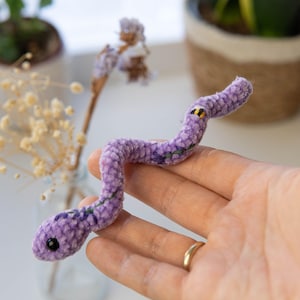 K&ouml;nnte beinhalten: Eine lila Pl&uuml;schschlange mit gestickten Details. Die Schlange ist in einer Hand gekr&uuml;mmt.