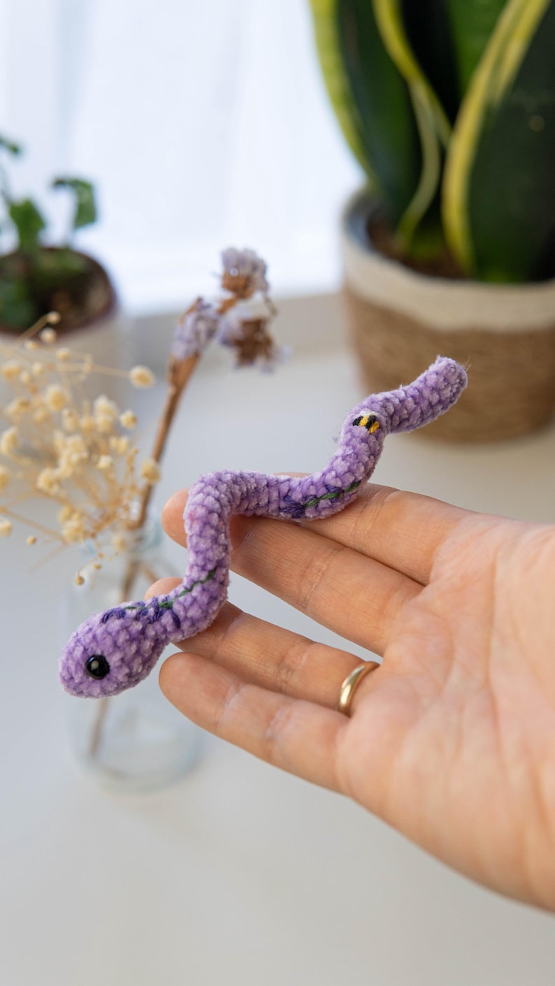 K&ouml;nnte beinhalten: Eine lila Pl&uuml;schschlange mit gestickten Details. Die Schlange ist in einer Hand gekr&uuml;mmt.