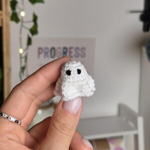 Puede incluir: Un peque&ntilde;o fantasma blanco tejido a mano con dos ojos negros. El fantasma se sostiene en una mano, con un fondo borroso que incluye la palabra "PROGRESS". Este art&iacute;culo es probablemente una pieza decorativa o un juguete peque&ntilde;o.