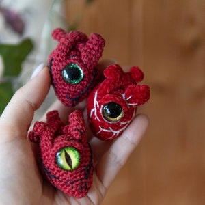 May include: Three crocheted red hearts with embroidered details and large googly eyes. The hearts are held in a hand.
