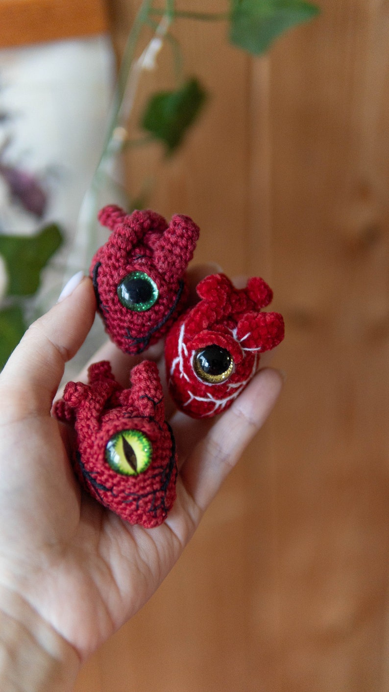 May include: Three crocheted red hearts with embroidered details and large googly eyes. The hearts are held in a hand.
