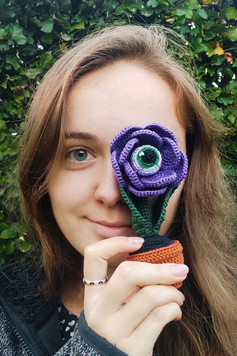 Puede incluir: Una flor de color morado hecha a crochet con un ojo verde en el centro. La flor se sostiene frente a la cara de una persona, cubriendo un ojo.