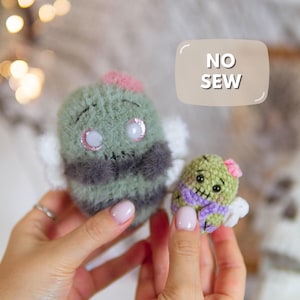 May include: Two fuzzy, rounded plush figures with stitched facial features and small wings. The larger figure is sage green with dark gray accents and a pink top, while the smaller one is light green with a purple scarf and pink flower.