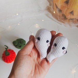 May include: Two white crocheted ghost ornaments with black eyes and pink cheeks. The ghosts are held in a hand against a white background.