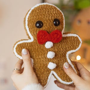 May include: A plush gingerbread man with a brown body, white icing trim, and a red bow tie. It has black button eyes and white buttons down the front. The handmade Christmas decoration is held by a person.
