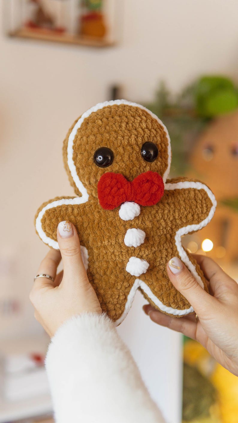 May include: A plush gingerbread man with a brown body, white icing trim, and a red bow tie. It has black button eyes and white buttons down the front. The handmade Christmas decoration is held by a person.