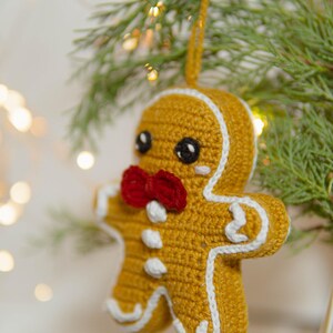 May include: A crocheted gingerbread man Christmas ornament. The ornament is yellow with white trim, a red bow tie, and black eyes. It is hanging from a green branch with small lights in the background.