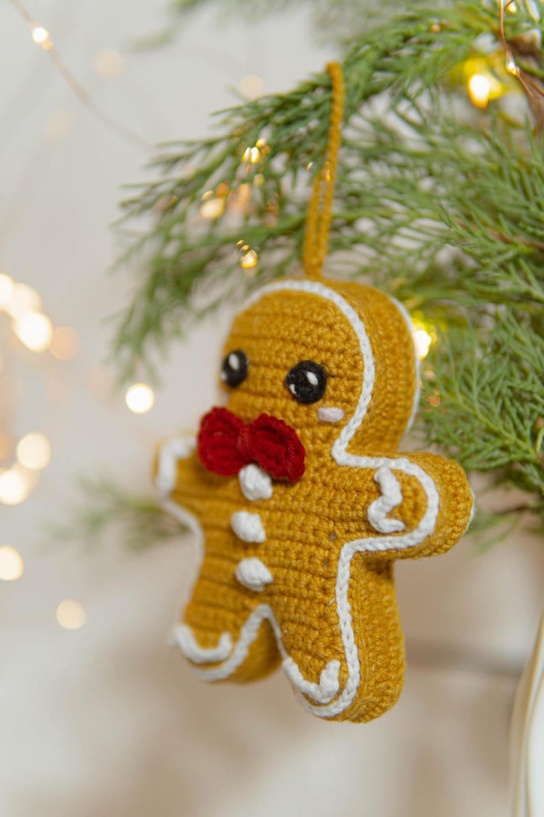 May include: A crocheted gingerbread man Christmas ornament. The ornament is yellow with white trim, a red bow tie, and black eyes. It is hanging from a green branch with small lights in the background.
