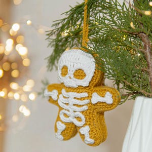 May include: A handmade, crochet gingerbread man ornament with a skeleton design. The ornament is yellow with white accents, including a skull and rib cage. It hangs from a yellow loop on a green tree branch.