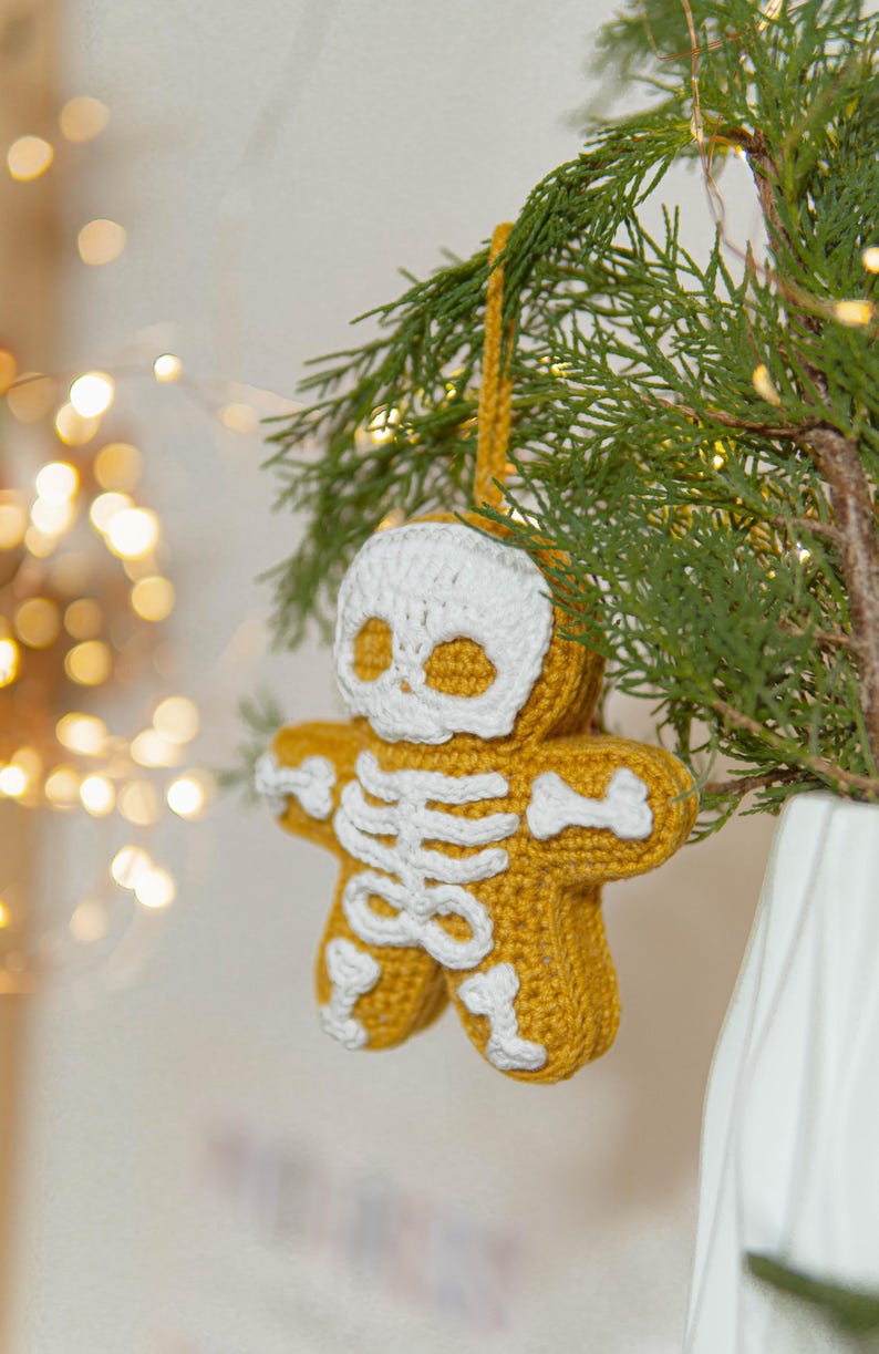May include: A handmade, crochet gingerbread man ornament with a skeleton design. The ornament is yellow with white accents, including a skull and rib cage. It hangs from a yellow loop on a green tree branch.