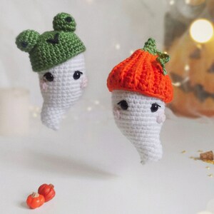 May include: Two crocheted ghost ornaments with different hats. One ghost wears a green frog hat and the other wears an orange pumpkin hat. The ghosts are on a white surface with small orange pumpkins.