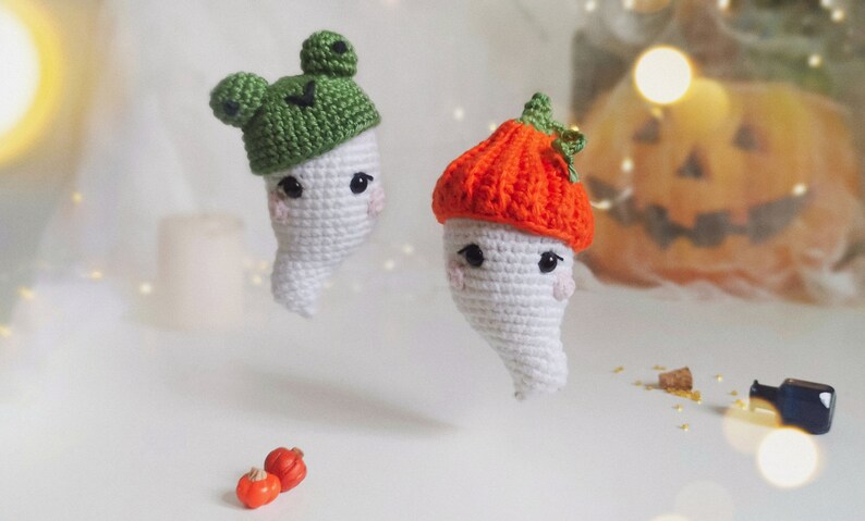 May include: Two crocheted ghost ornaments with different hats. One ghost wears a green frog hat and the other wears an orange pumpkin hat. The ghosts are on a white surface with small orange pumpkins.