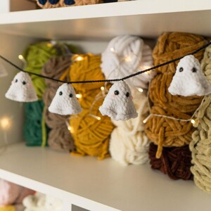 Puede incluir: Una guirnalda tem&aacute;tica de Halloween con adornos blancos en forma de fantasma con ojos negros, ensartados en un cord&oacute;n negro. La guirnalda se exhibe frente a varios ovillos de hilo de colores, incluyendo amarillo, marr&oacute;n y verde. Las luces de hadas a&ntilde;aden un brillo c&aacute;lido.