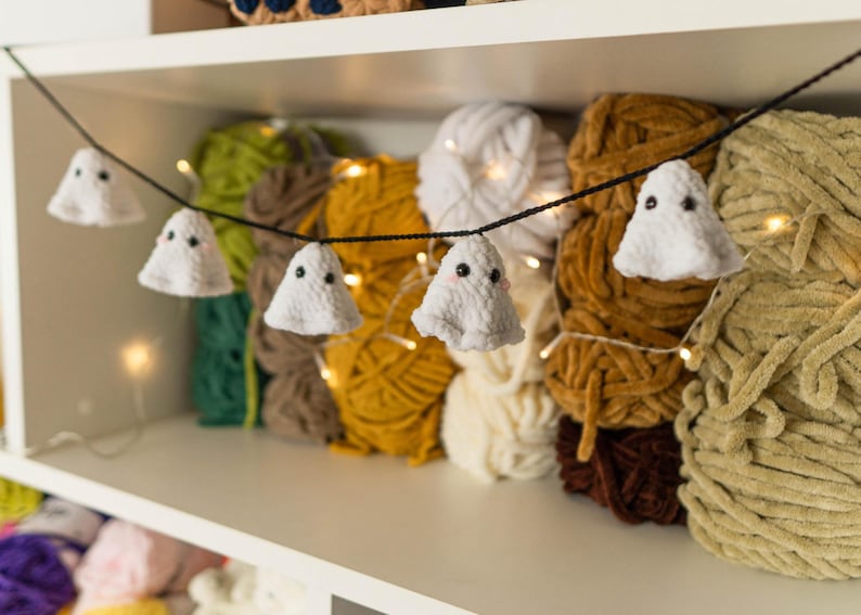 Puede incluir: Una guirnalda tem&aacute;tica de Halloween con adornos blancos en forma de fantasma con ojos negros, ensartados en un cord&oacute;n negro. La guirnalda se exhibe frente a varios ovillos de hilo de colores, incluyendo amarillo, marr&oacute;n y verde. Las luces de hadas a&ntilde;aden un brillo c&aacute;lido.