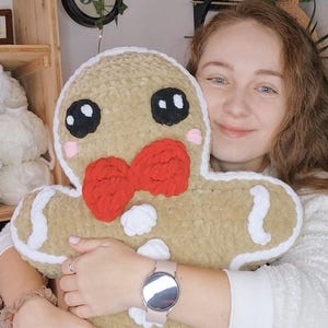 May include: A large, plush gingerbread man pillow with a tan body, white frosting trim, and a red bow tie. The pillow has black eyes, pink cheeks, and a white button. The pillow is soft and cuddly.