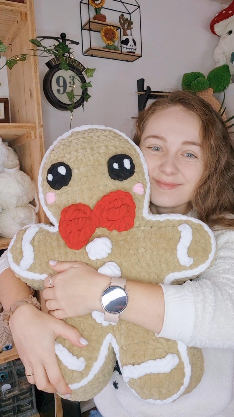 May include: A large, plush gingerbread man pillow with a tan body, white frosting trim, and a red bow tie. The pillow has black eyes, pink cheeks, and a white button. The pillow is soft and cuddly.