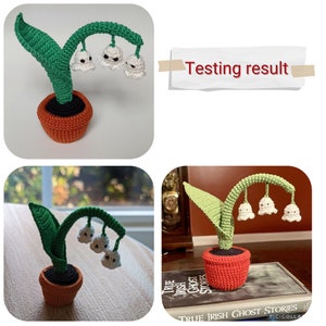 May include: Three crocheted white lily of the valley flowers with black eyes hang from a green stem. The stem is attached to a crocheted orange pot with a black rim.