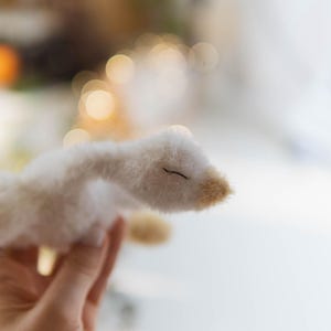 May include: A handmade white plush goose with a beige beak and closed eyes. The soft toy is held in a hand, with a blurred background of lights and a window. The goose is a whimsical, handcrafted item.