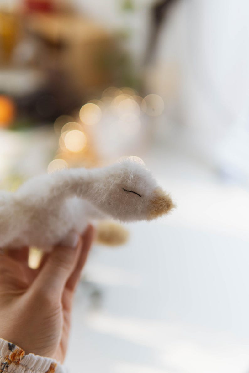May include: A handmade white plush goose with a beige beak and closed eyes. The soft toy is held in a hand, with a blurred background of lights and a window. The goose is a whimsical, handcrafted item.