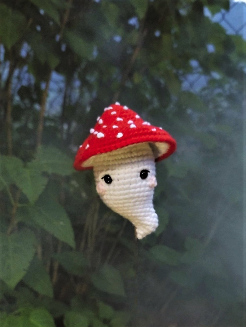 May include: A crocheted mushroom toy with a red and white cap and a white body. The mushroom has a cute face with black eyes and a small smile.