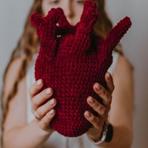 May include: A person holding a large, red crocheted heart. The heart is made of yarn and has a textured surface.