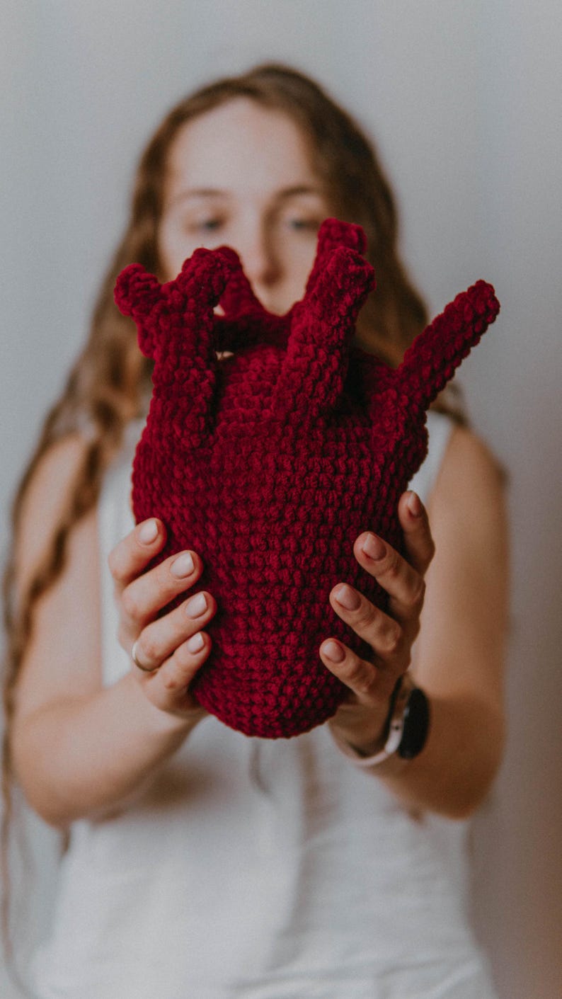 May include: A person holding a large, red crocheted heart. The heart is made of yarn and has a textured surface.