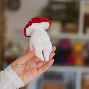 以下が含まれることがあります： 白と赤のドット柄の傘を持つ手作りの編み物キノコのおもちゃ。キノコは手で持たれており、柔らかい質感と気まぐれなデザインを強調しています。保育園の装飾に最適です。