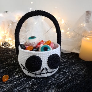 K&ouml;nnte beinhalten: Ein wei&szlig;er geh&auml;kelter Halloween-S&uuml;&szlig;igkeitenkorb in Form eines Sch&auml;dels mit schwarzen Augen und einem gestickten Mund. Der Korb ist gef&uuml;llt mit S&uuml;&szlig;igkeiten und Spielzeugaugen.