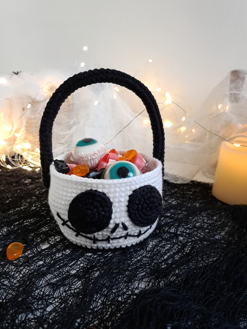 K&ouml;nnte beinhalten: Ein wei&szlig;er geh&auml;kelter Halloween-S&uuml;&szlig;igkeitenkorb in Form eines Sch&auml;dels mit schwarzen Augen und einem gestickten Mund. Der Korb ist gef&uuml;llt mit S&uuml;&szlig;igkeiten und Spielzeugaugen.