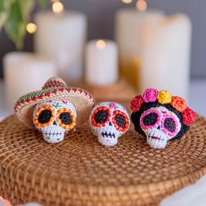 May include: Three crocheted sugar skull figurines. The left skull wears a tan sombrero with green and red trim. The center skull is smaller with orange and black eye sockets. The right skull features a black headpiece with pink, yellow, and orange flowers.