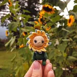 K&ouml;nnte beinhalten: Eine geh&auml;kelte Sonnenblumenpuppe mit gr&uuml;nem K&ouml;rper und gelben und blauen Bl&uuml;tenbl&auml;ttern. Die Puppe wird in einer Hand mit einem silbernen Ring gehalten.