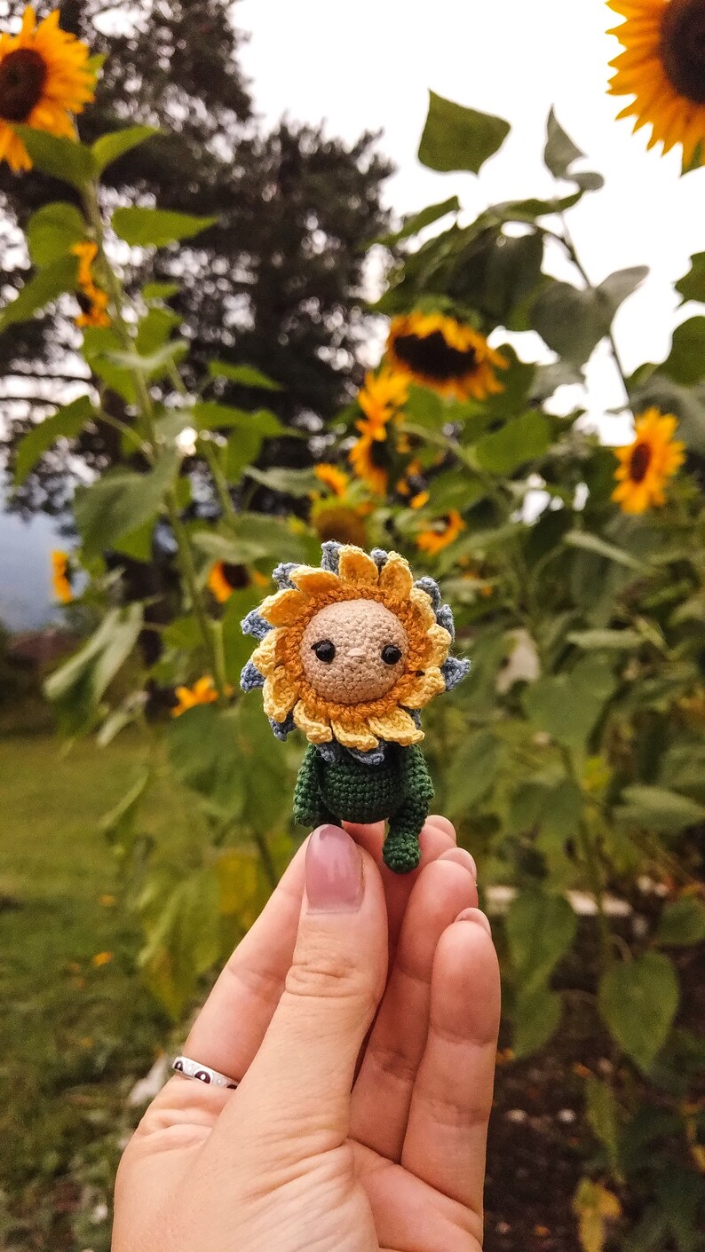 K&ouml;nnte beinhalten: Eine geh&auml;kelte Sonnenblumenpuppe mit gr&uuml;nem K&ouml;rper und gelben und blauen Bl&uuml;tenbl&auml;ttern. Die Puppe wird in einer Hand mit einem silbernen Ring gehalten.