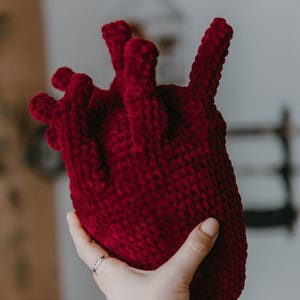 May include: A crocheted heart-shaped plush toy in a deep red color. The toy is held in a hand, showcasing its detailed texture and three-dimensional form.