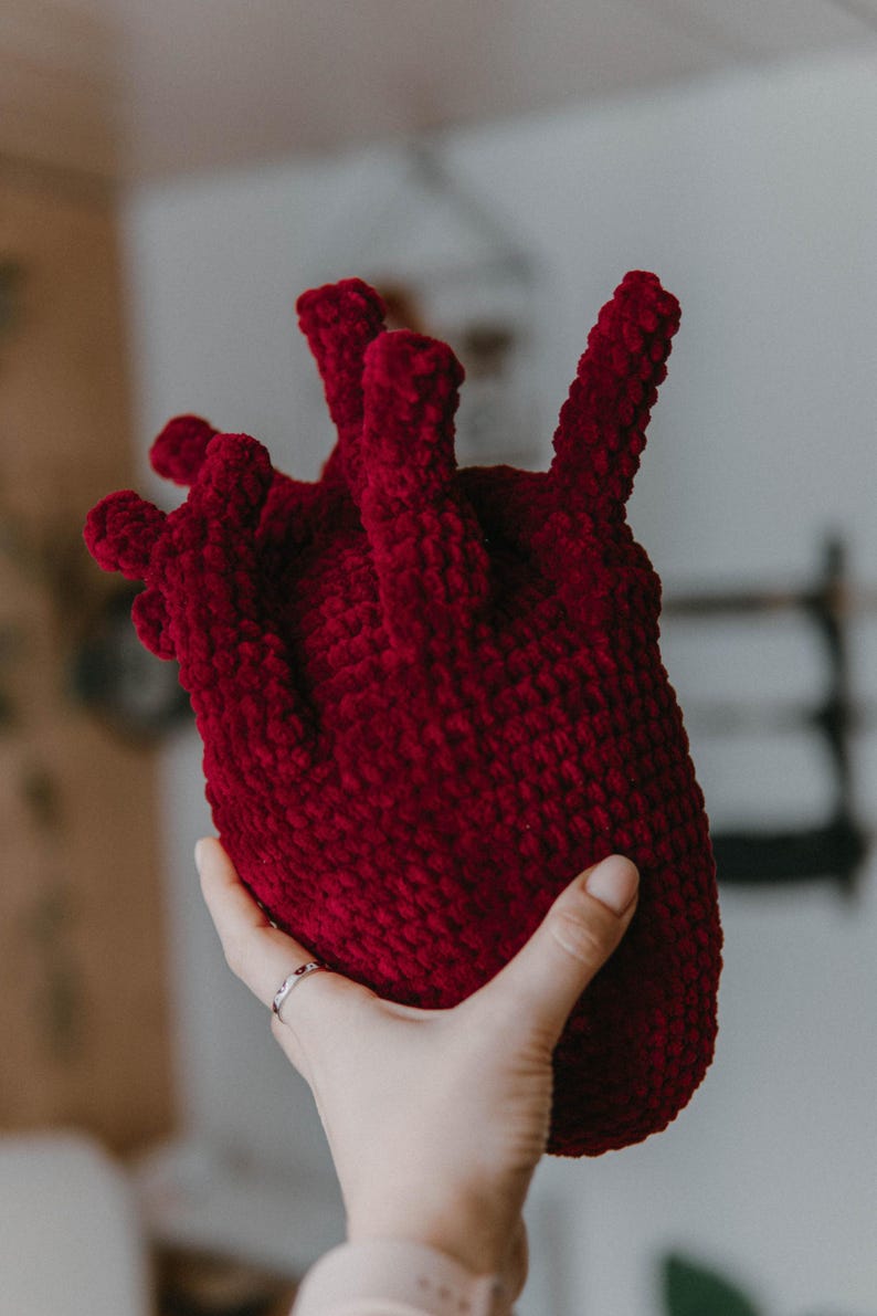 May include: A crocheted heart-shaped plush toy in a deep red color. The toy is held in a hand, showcasing its detailed texture and three-dimensional form.