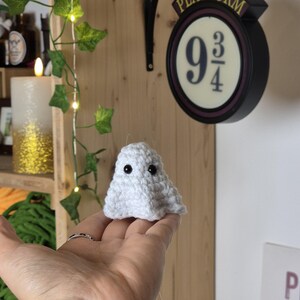 Puede incluir: Un peque&ntilde;o fantasma blanco de ganchillo hecho a mano con dos ojos negros. El fantasma est&aacute; sostenido en la mano de una persona. El fondo incluye un letrero que dice "Platform 9 3/4".