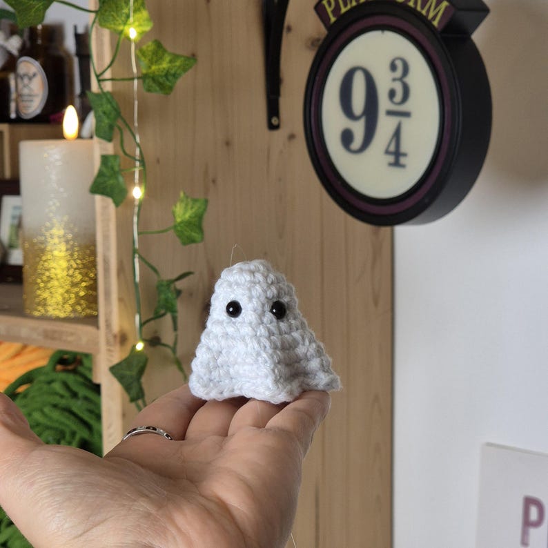 Puede incluir: Un peque&ntilde;o fantasma blanco de ganchillo hecho a mano con dos ojos negros. El fantasma est&aacute; sostenido en la mano de una persona. El fondo incluye un letrero que dice "Platform 9 3/4".
