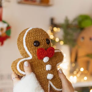 May include: A plush gingerbread man with a brown body, white icing details, and a red bow tie. The gingerbread man has black button eyes and white buttons. The plush toy is held in front of a blurred background with Christmas decorations.