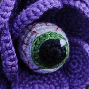 Puede incluir: Una flor de crochet morado con un ojo blanco, un iris verde y venas rojas.
