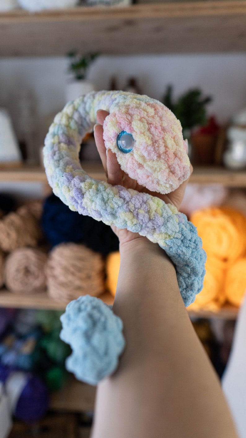 K&ouml;nnte beinhalten: Ein geh&auml;keltes Schlangenspielzeug mit einem Pastellfarbschema, einschlie&szlig;lich Rosa, Gelb, Blau und Lila. Die Schlange hat ein einzelnes blaues Auge und ist zu einer Schleife gekr&auml;uselt.