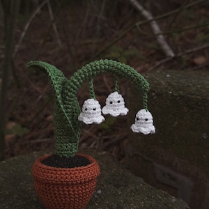 May include: A crocheted lily of the valley plant in a brown pot. The plant has white flowers with black eyes and green leaves.