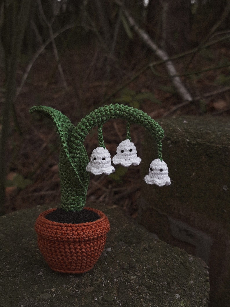 May include: A crocheted lily of the valley plant in a brown pot. The plant has white flowers with black eyes and green leaves.