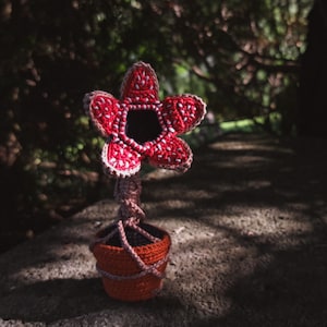May include: A crocheted red and white flower with a black center, in a brown pot. The flower is a replica of the plant from the movie Stranger Things.