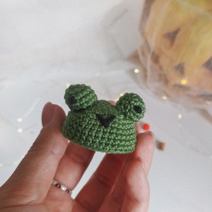 May include: A small green crocheted frog with black eyes. The frog is being held by a hand.
