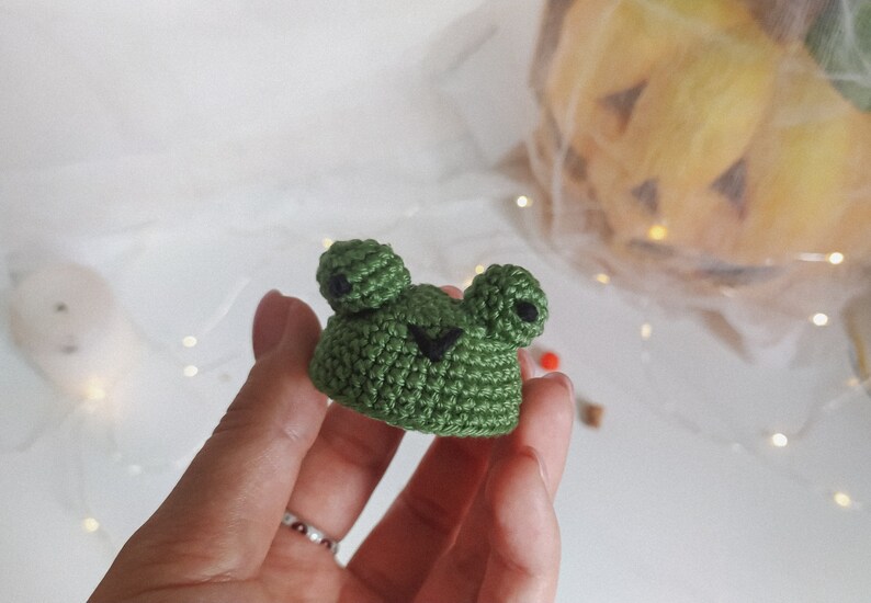 May include: A small green crocheted frog with black eyes. The frog is being held by a hand.