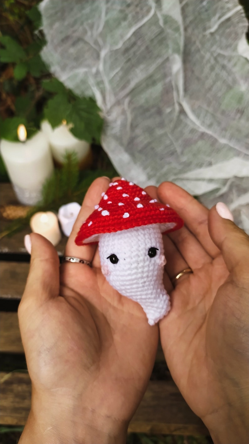 May include: A crocheted mushroom toy with a red and white cap and a white body. The mushroom has black eyes and a cute expression. The toy is being held in a person's hands.