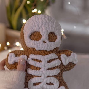 May include: A brown crocheted gingerbread man with a white skull face and skeleton design. The plush toy is handmade and has a soft, textured appearance. The background is blurred, with hints of greenery and fairy lights.